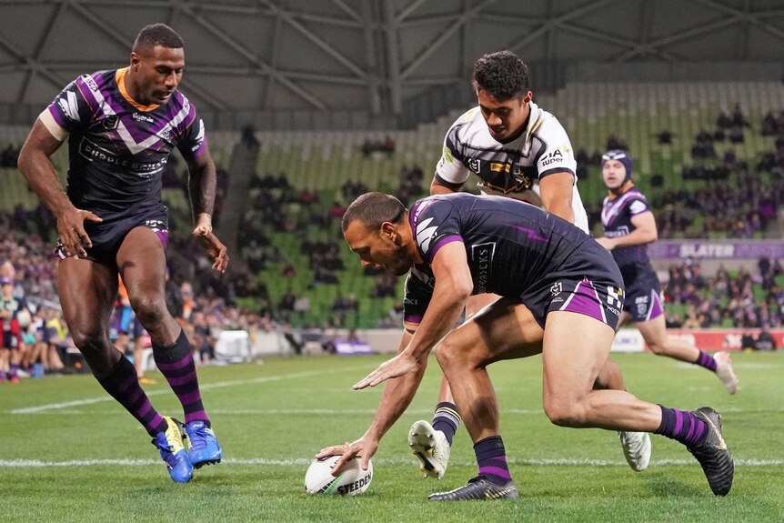 An NRL player plants the ball down over the try line as a teammate and opponent watch on.
