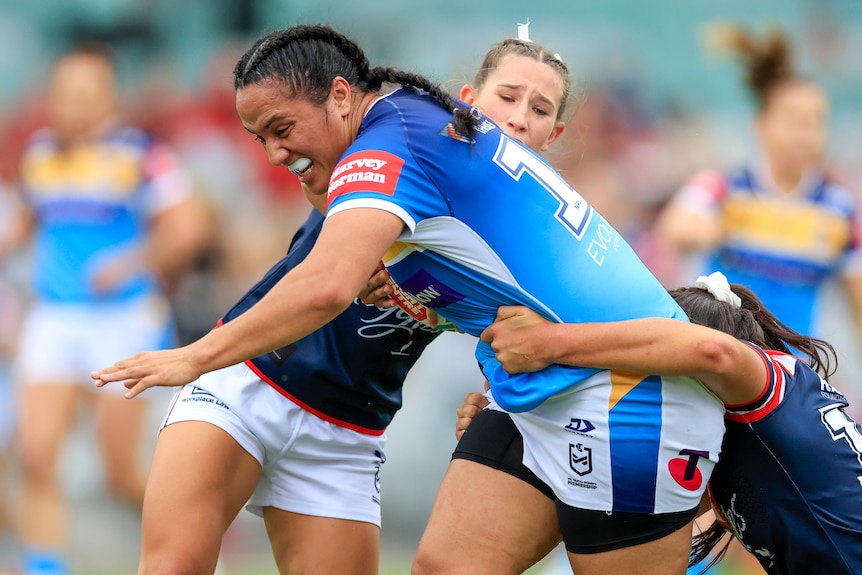 A Gold Coast Titans NRLW player carries the ball while being tackled by Sydney Roosters opponents.