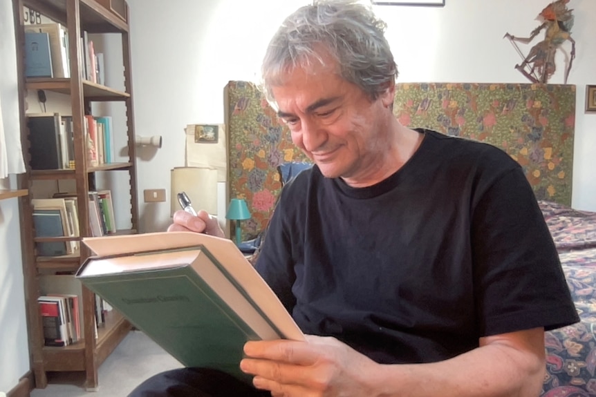Carlo Rovelli sitting and signing sheet of paper, leaning on book. 