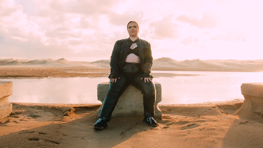 Djanaba, a young woman wearing a black bra under a black jacket and black pants, sits in front of a body of water near dunes.
