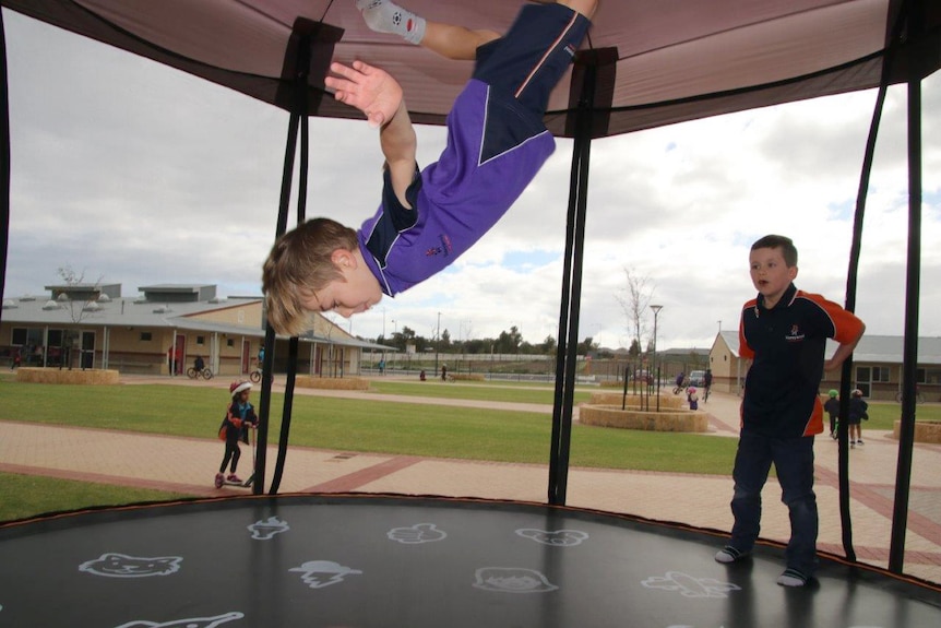 A student wearing a purple shirt does a somersault on a trampoline as another child watches on.