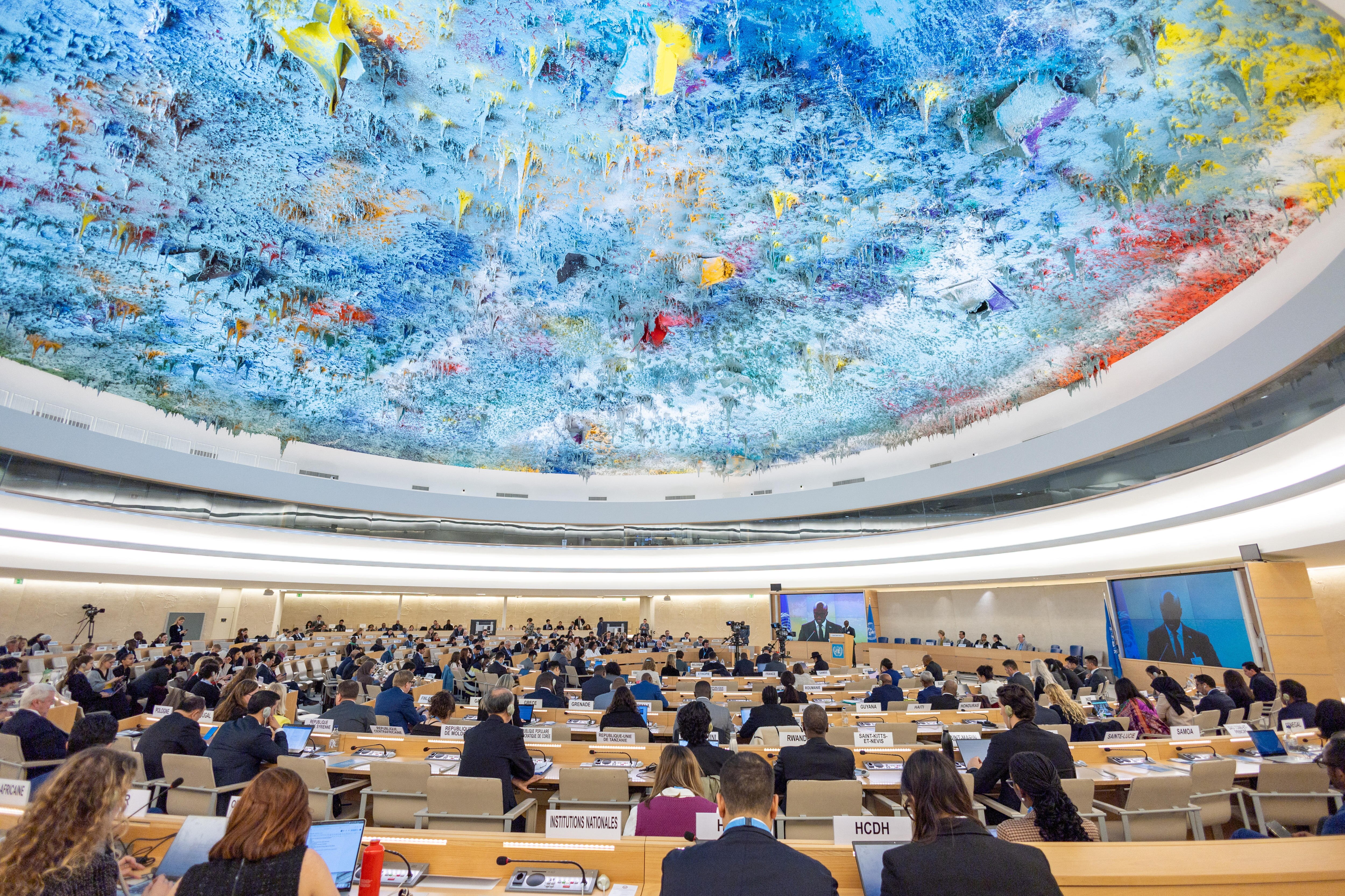 A wideshot of a debate hall, with lots of seats and diplomats sitting in them.