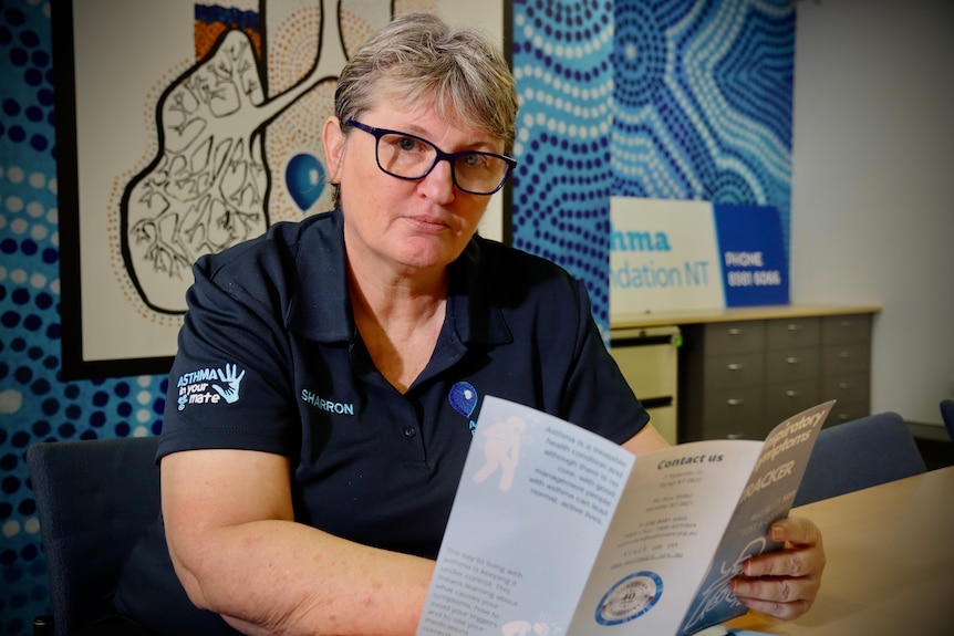 A middle-aged woman with short hair sits at a table holding a pamphlet and looking at the camera