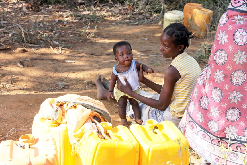 A woman waits with her baby to collect water from a hole in the ground.