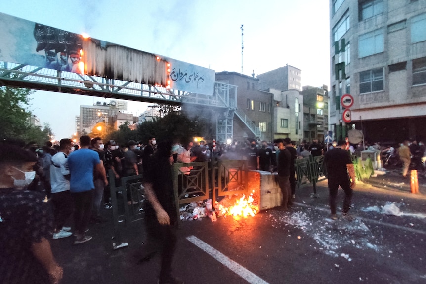 A mass of young men gather around a makeshift blockade on a Tehran street as a fire burns at their feet.