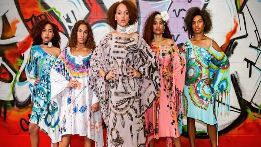 Five pacific islands women with dark brown afro hair wearing blue, pink, brown and green dresses in front of a graffiti wall