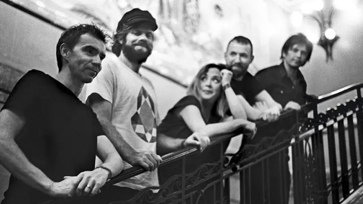 Five members of the band Slowdive standing on a staircase, looking over the railing and smiling.