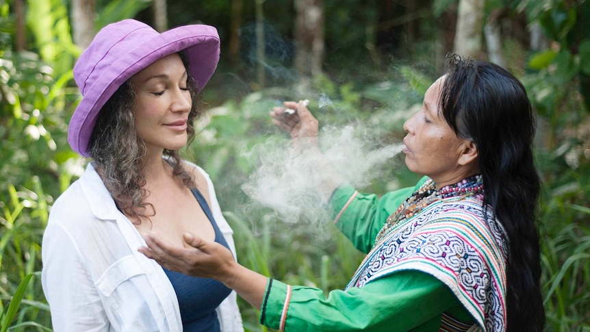 A female shaman exhales a plume of smoke over a white woman in a hat