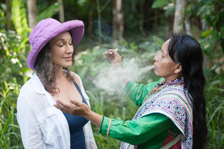 A female shaman exhales a plume of smoke over a white woman in a hat