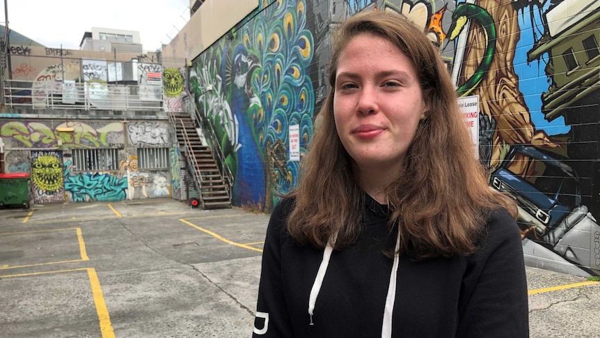 A young woman stands in front of a graffitied wall.