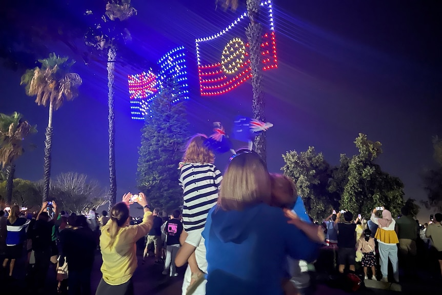 A light show illuminates flags in the Perth sky at night on Australia Day, with people watching from the ground.