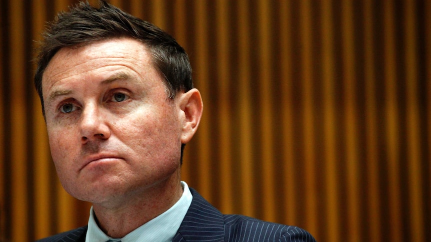 A man with short, dark hair, wearing a dark suit, apparently listening to something with a serious look on his face.