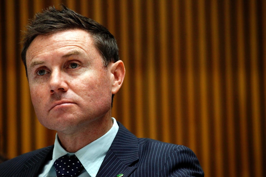 A man with short, dark hair, wearing a dark suit, apparently listening to something with a serious look on his face.