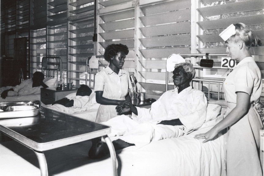 A black and white photograph of a patient in bed with nurses.