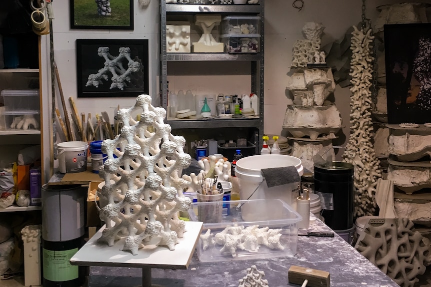 A crowded industrial design studio is covered with prototypes on walls and plinths, with a dust-covered desk in the foreground.