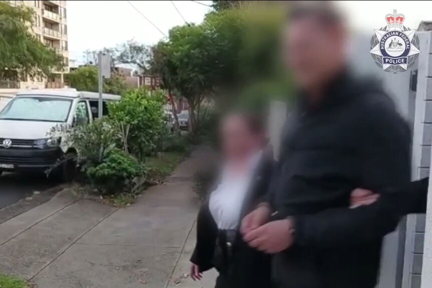 two female police officers hold onto the arm of a man in handcuffs as they walk him out of a house