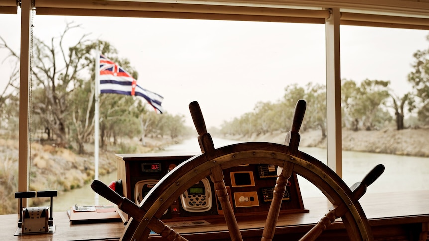 The wooden wheel of a paddle steamer with the boat's dashboard and a river in the background.