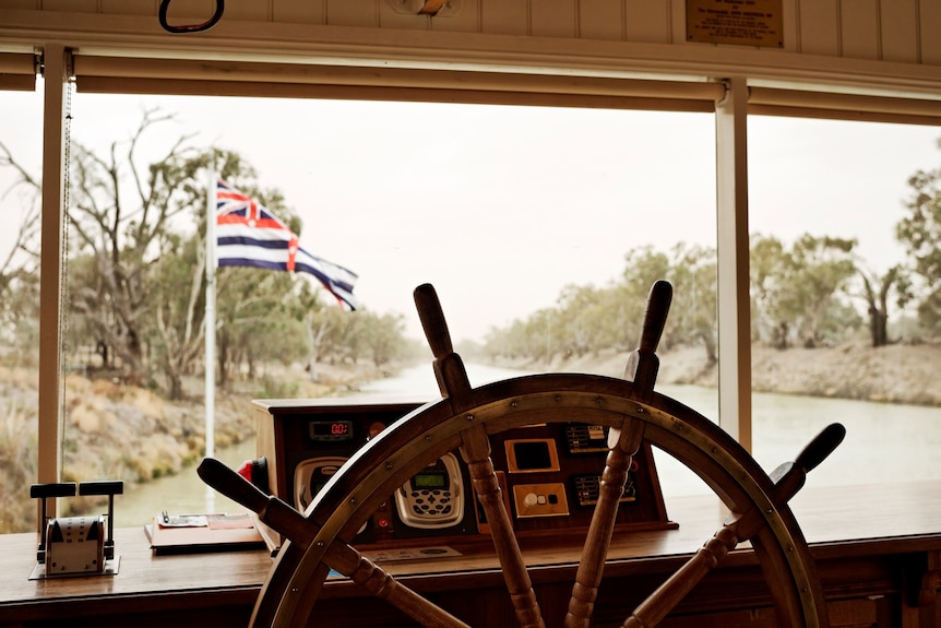 The wooden wheel of a paddle steamer with the boat's dashboard and a river in the background.