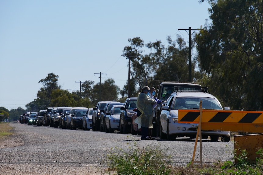 A queue of cars with medical professionals in COVID protective gear at the front of the queue.