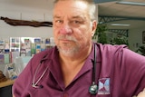 close up of man leaning on a clinic reception counter wearing a purple work shirt and a stethoscope