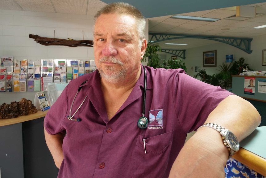 close up of man leaning on a clinic reception counter wearing a purple work shirt and a stethoscope