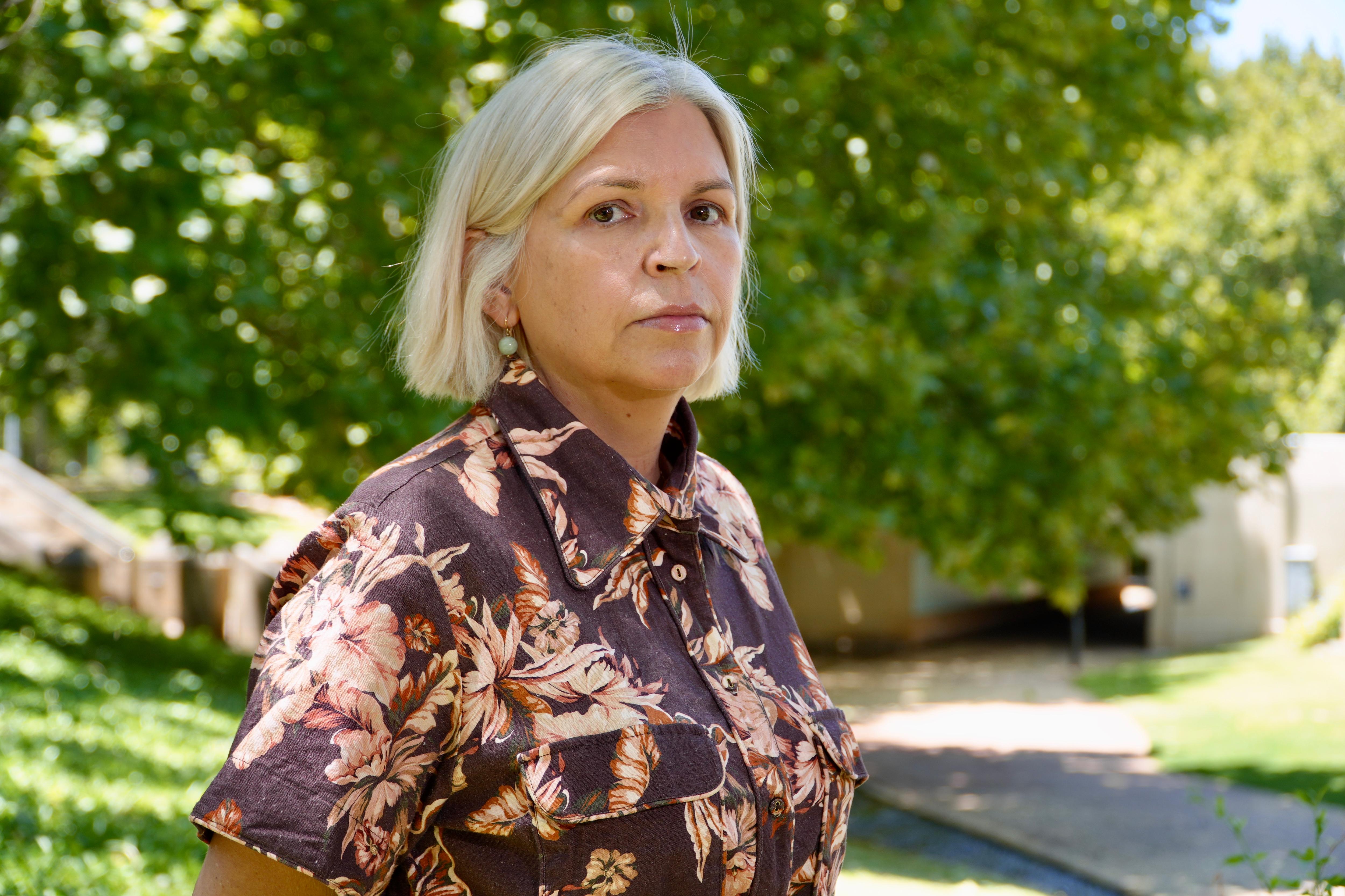 Una mujer con cabello rubio lacio viste un traje estampado. Ella está parada afuera y tiene una expresión seria en su rostro.