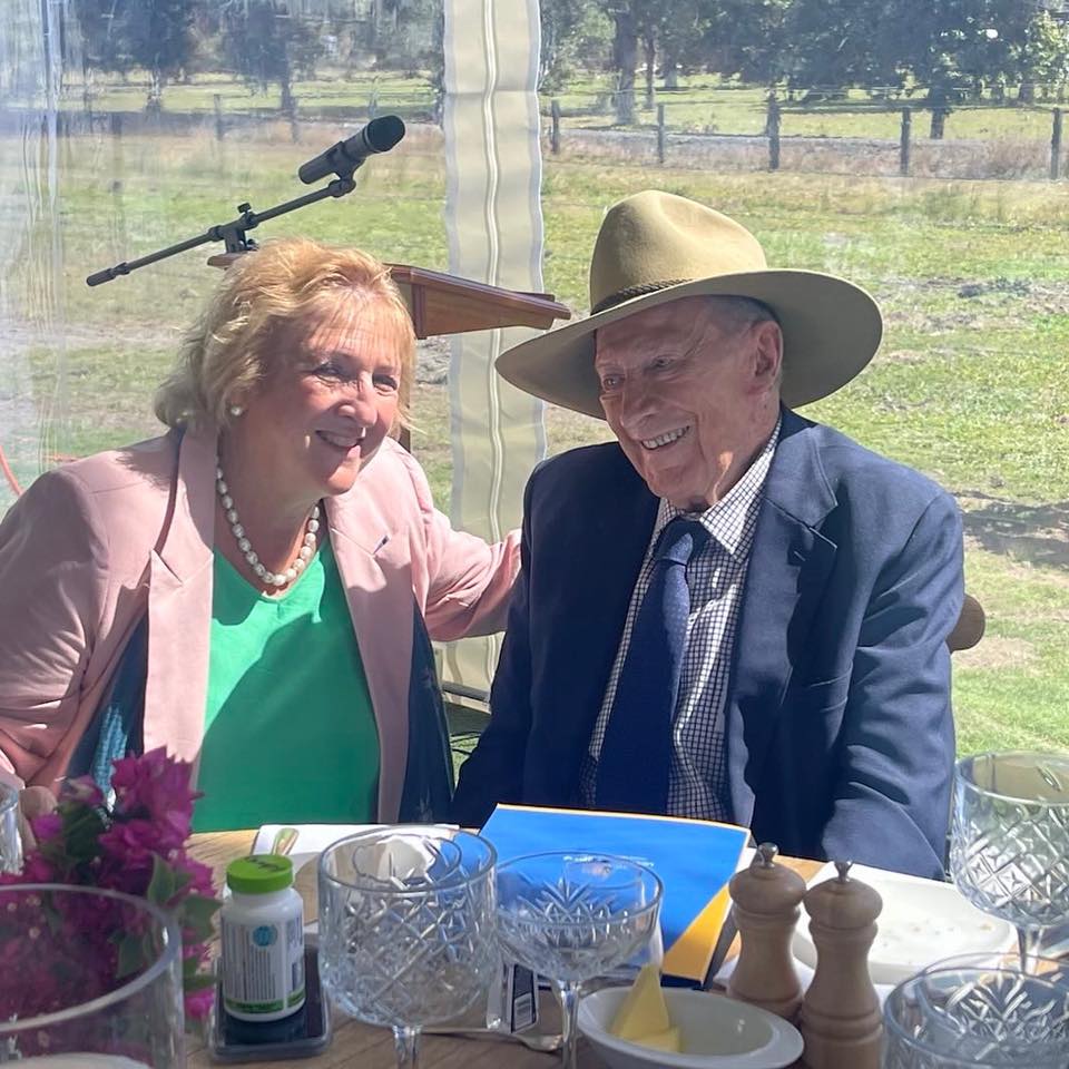 Una mujer con una chaqueta rosa sonríe sentada junto a un hombre mayor con un traje azul y un sombrero estilo Akubra.