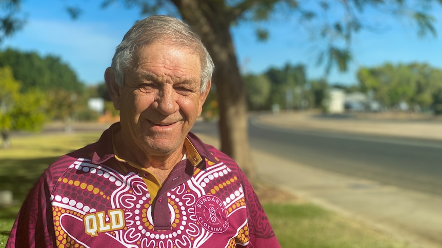 Head and shoulder mid shot portrait of an indigenous man in a maroons supporter shirt alongside a road