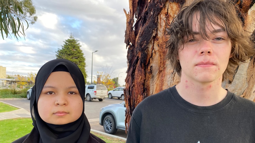 A young woman looks at camera, she wears black headscarf, young man has grown out brown hair, dark shirt. they are solemn