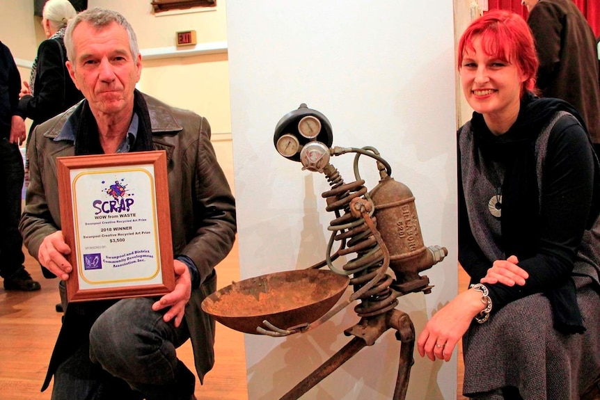 A man holding a winner's certificate and a woman crouch down to pose next to a sculpture of a tin man made of scrap metals.