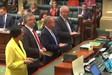 A woman wearing a yellow jacket and three men wearing dark suits stand at a table in parliament