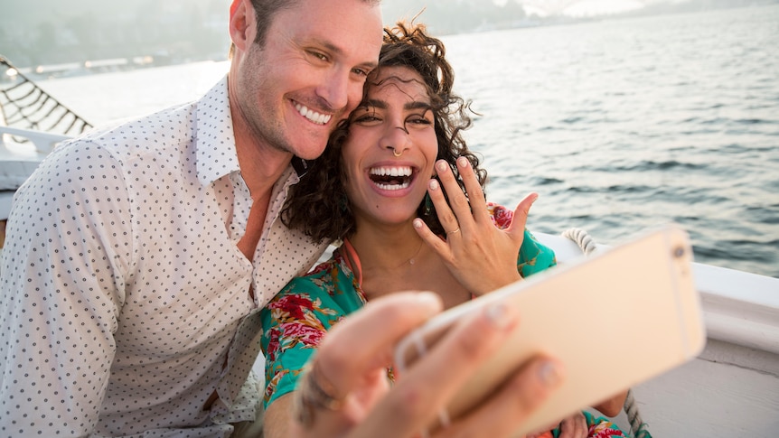 Close up of young smiling couple on a boat, girl holding out her hand with ring and taking selfie