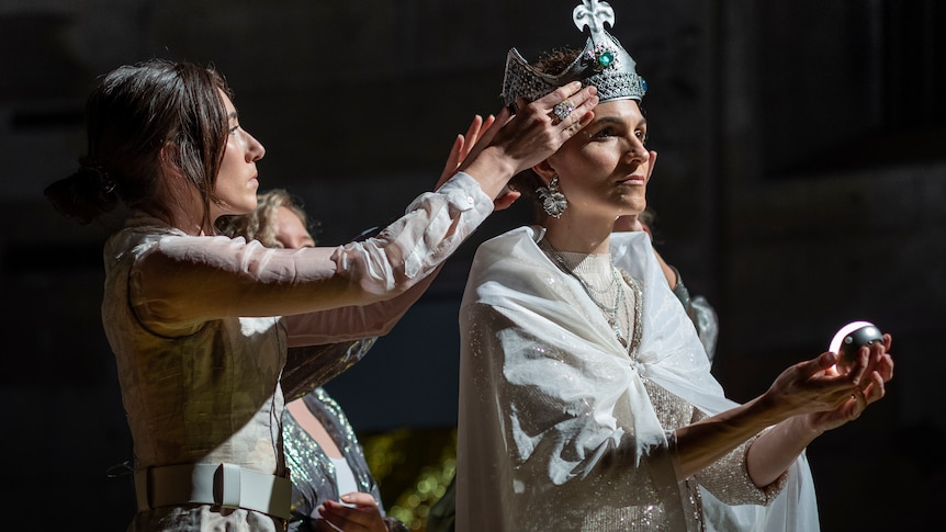 A singer wears white medieval robes on stage while another singer places a silver crown on her head.
