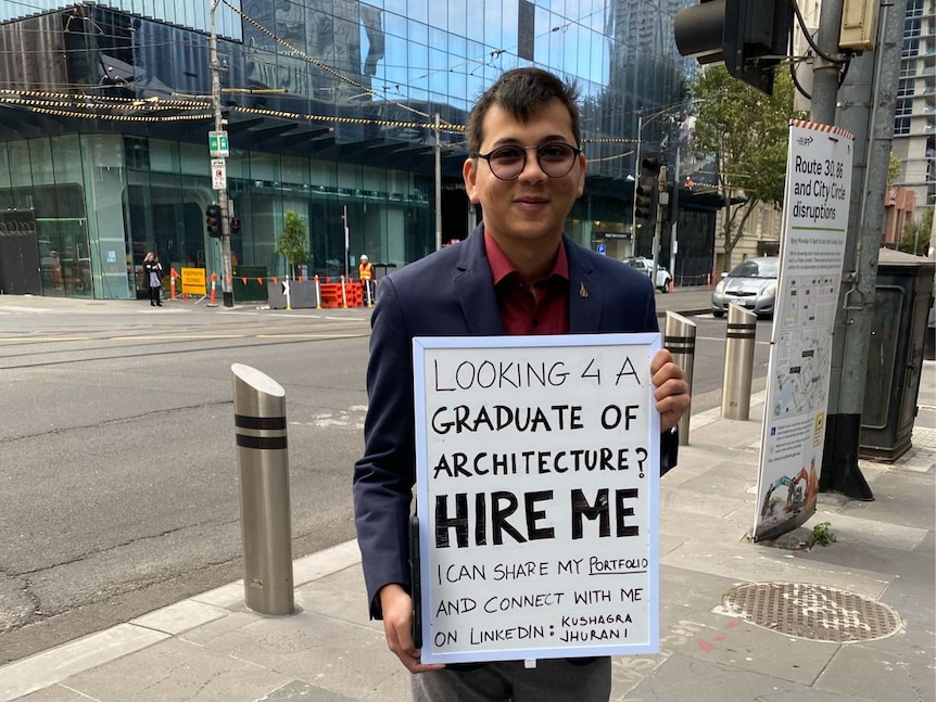 A young man dressed in a suit stands with a sing that reads looking 4 a graduate of architecture HIRE ME?