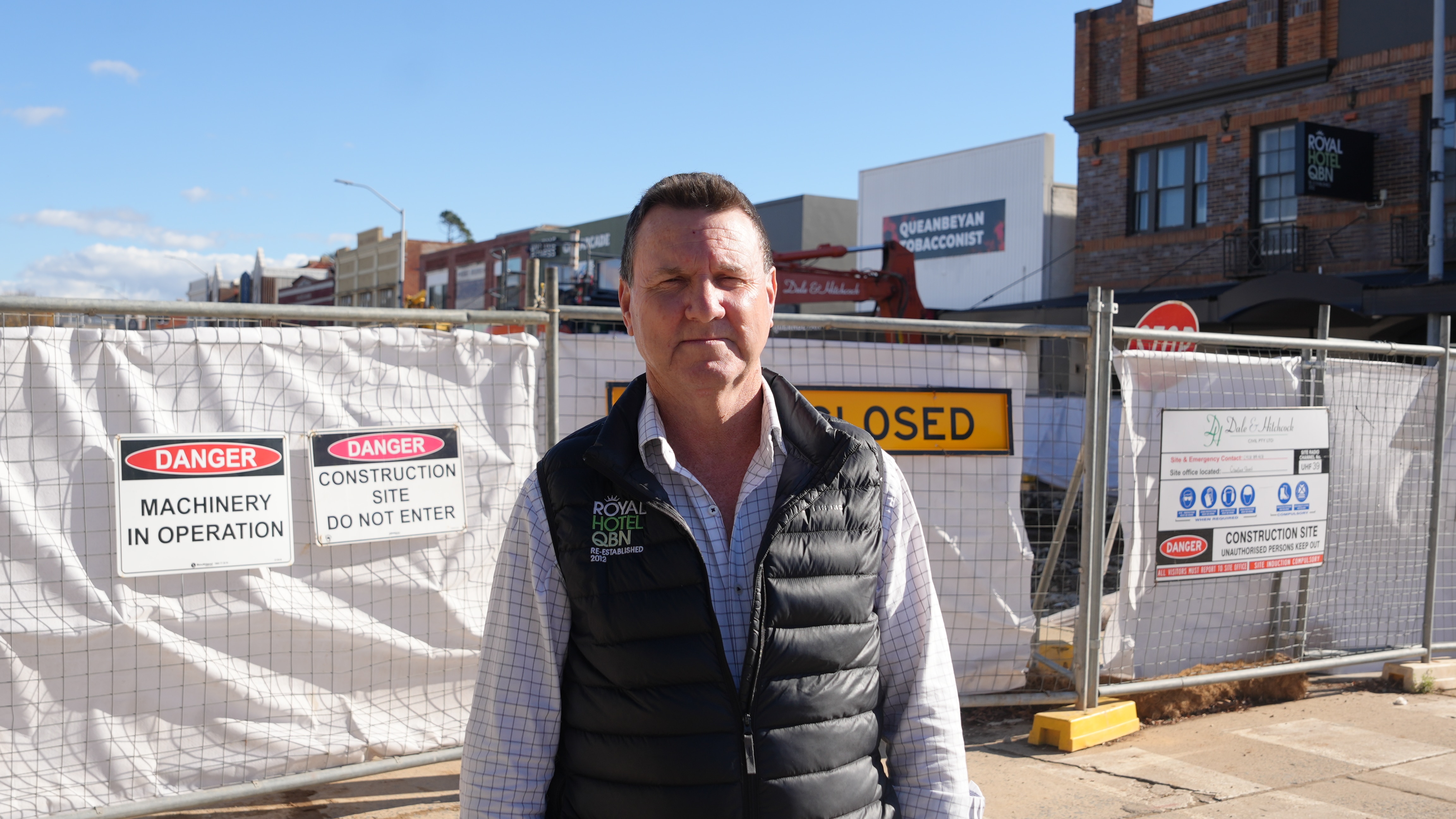 A man with short hair, wearing a shirt and puffer vest, stands outside in front of a closed road.