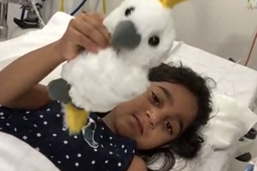 Tharnicaa lying in bed looking sad, holding stuffed toy cockatoo.