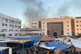 smoke rises above hospital building, while in the foreground makeshift tents can be seen