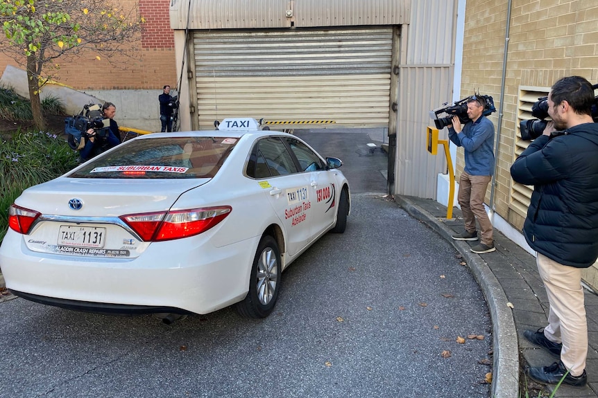 A taxi surrounded by media camera crews leaves an Adelaide court.