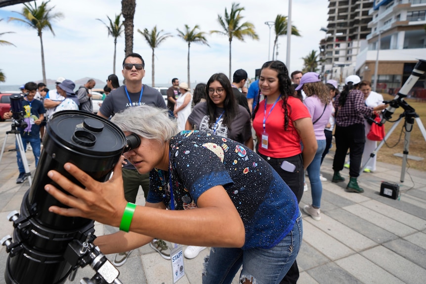 A woman sets up a telescope as a crowd watches on with palm trees in the background