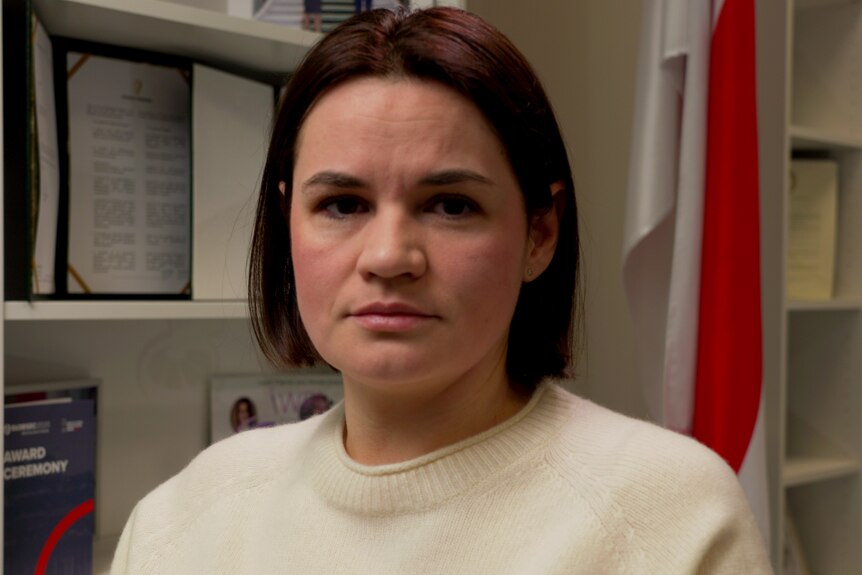 A woman with a short brown haircut and wearing a white jumper looks at the camera with a neutral expression