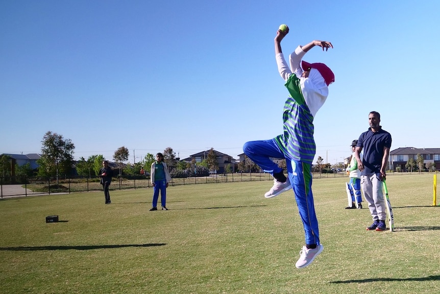 A girl dressed in a blue and green cricket tracksuit, hijab and cap leaps into the air as she prepares to bowl on an oval.