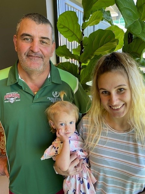 A man in a green shirt standing beside a smiling blonde woman who is holding a baby.
