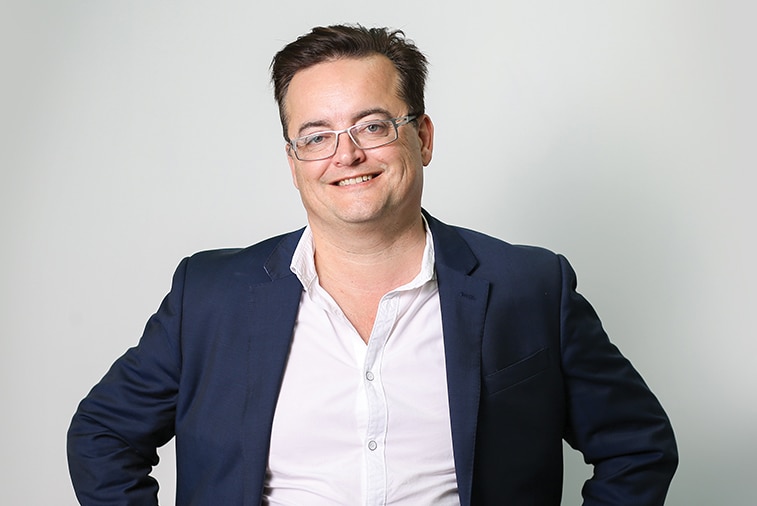 Man in suit and white shirt smiles, wearing grey rimmed glasses