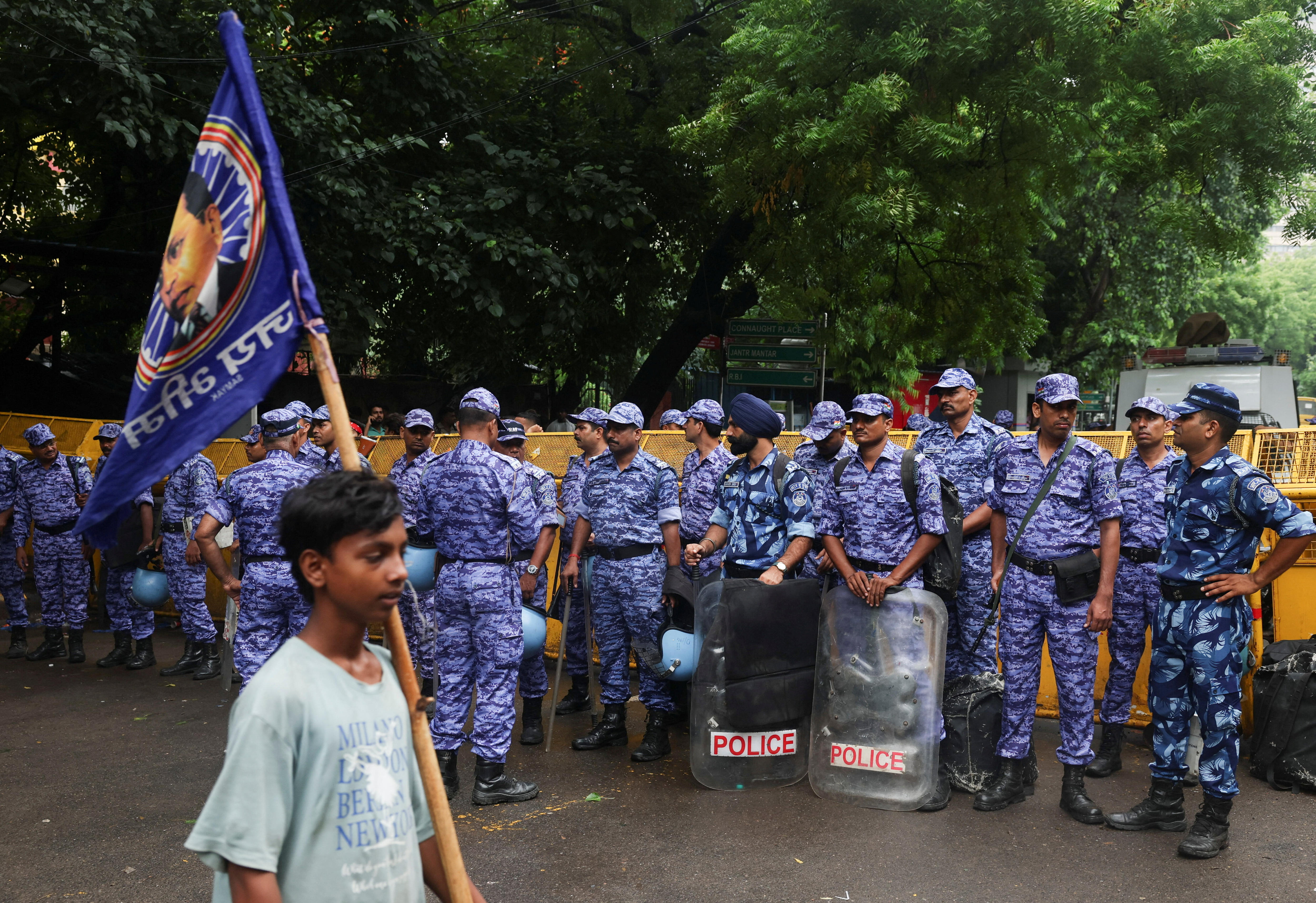 A man carrying a flag walks past a group of police in uniform holding shields. 
