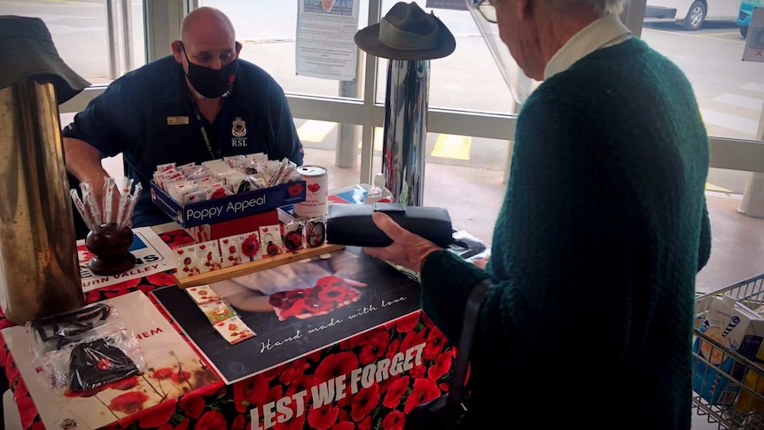 A woman hands over money to a man seated at a desk who is selling poppies and pins for fundraising, for Remembrance Day