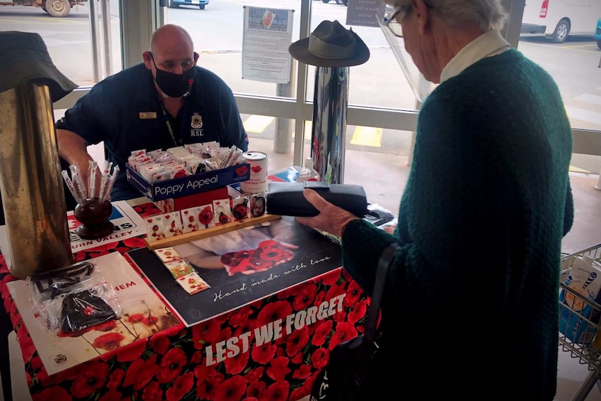 A woman hands over money to a man seated at a desk who is selling poppies and pins for fundraising, for Remembrance Day