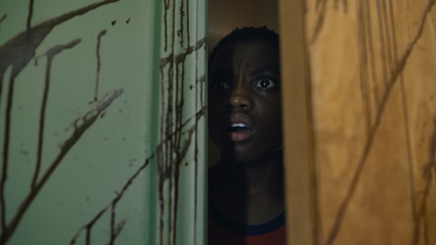 A close up of Rodney L Jones looking on in horror through a crack in a blood-spattered door.