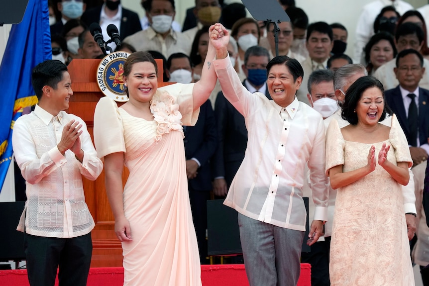 A smiling woman and man in formal clothes hold hands aloft as they celebrate before a large crowd.