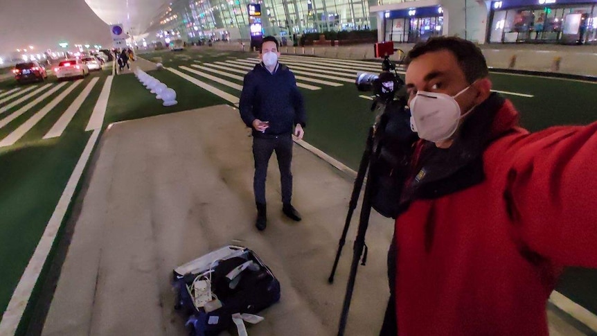 Cameraman filming journalist outside airport with both wearing face masks.