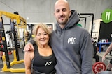 An older woman with blonde hair stands with arm around waist of taller man in hoodie in gym
